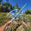 Person holding a blue fantasy weapon in a garden with trees and flowers
