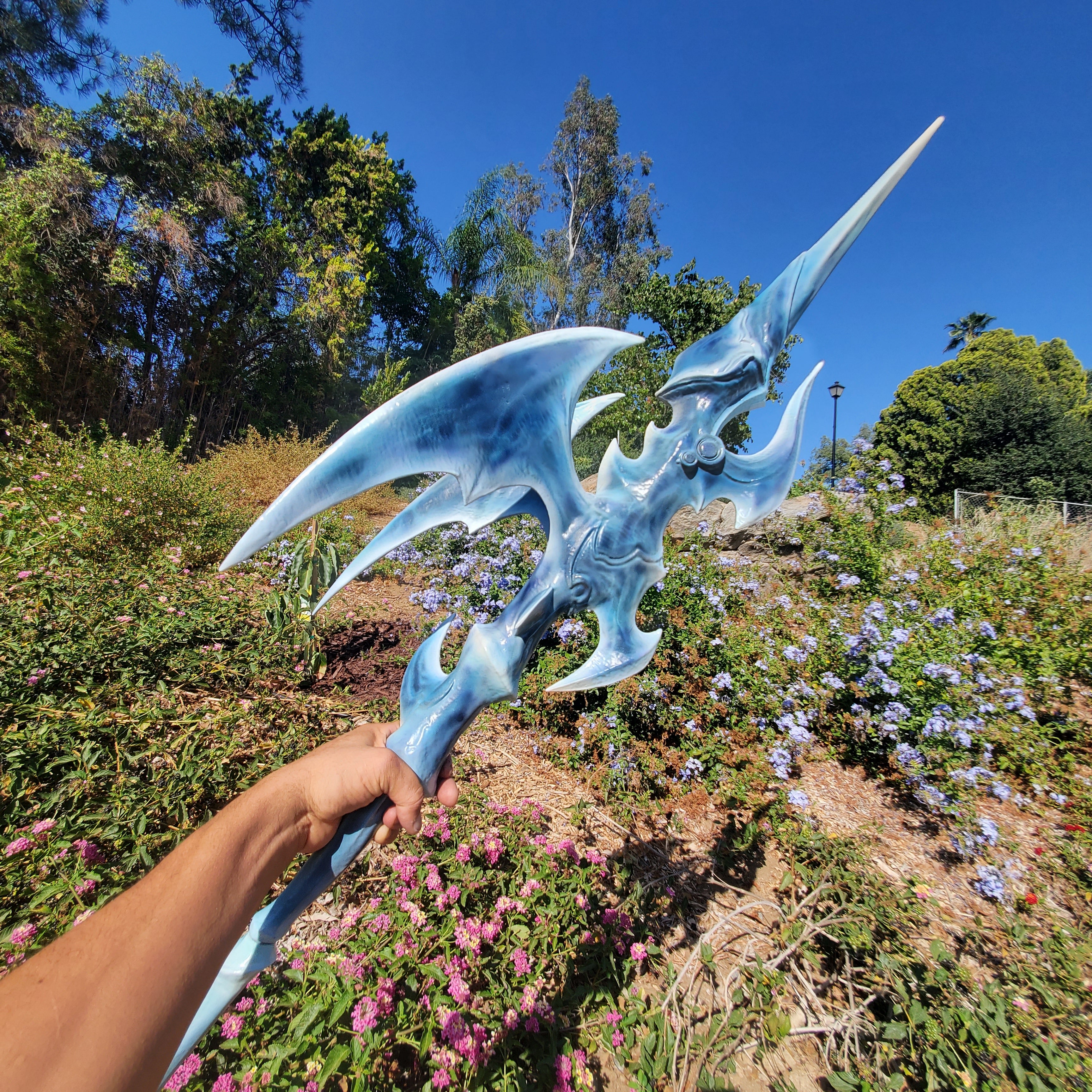 Person holding a blue fantasy weapon in a garden with trees and flowers