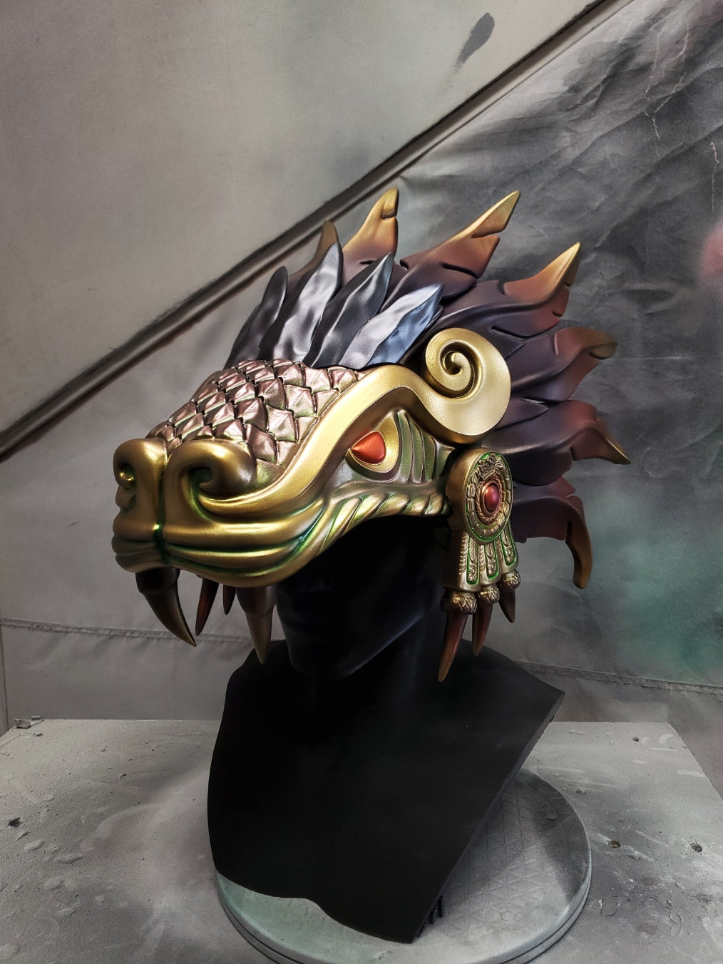 Hand‑sculpted Quetzalcóatl warrior mask with metallic gold, green, and violet detailing displayed on a black mannequin head