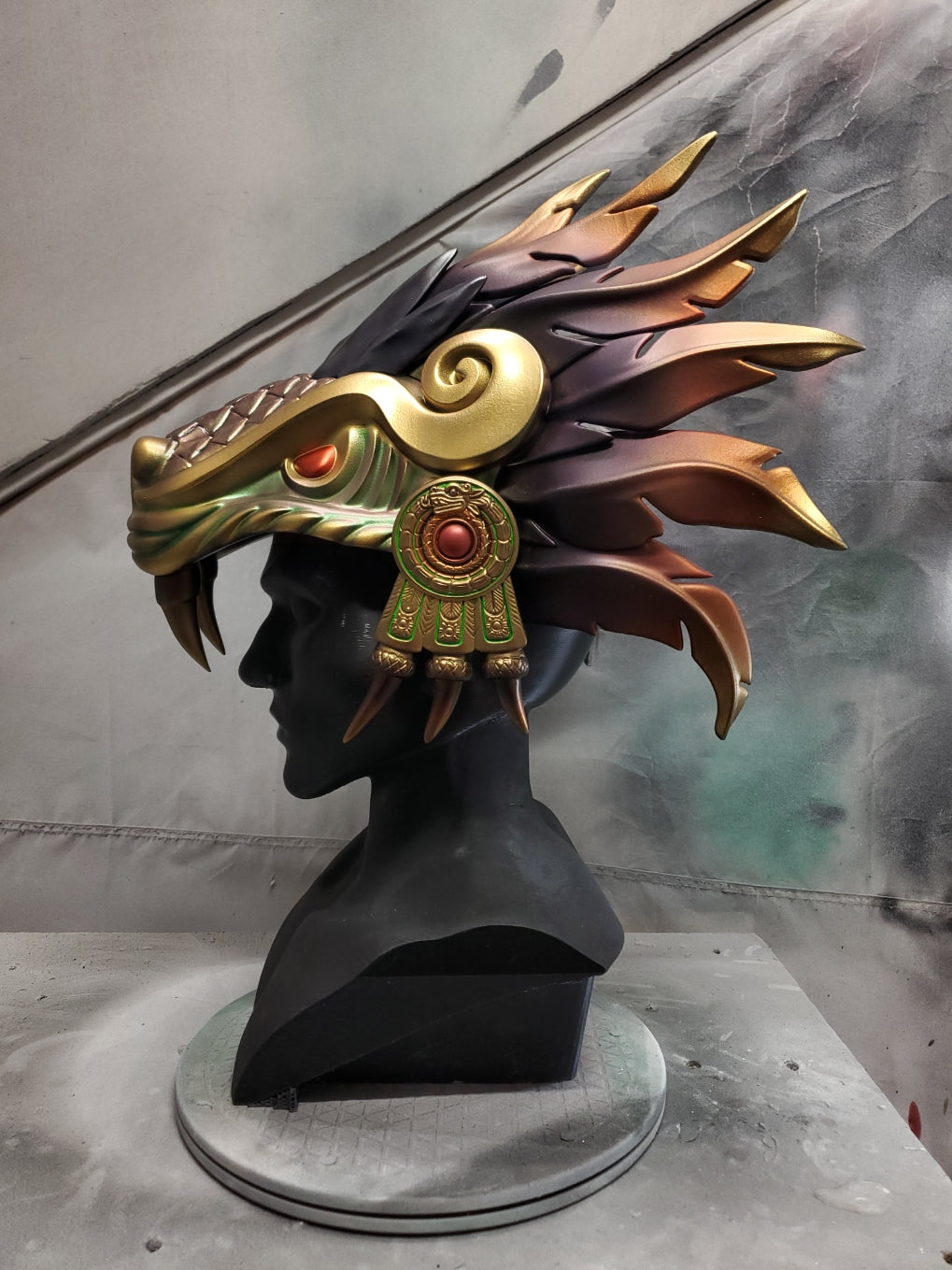 Hand‑sculpted Quetzalcóatl warrior mask with metallic gold, green, and violet detailing displayed on a black mannequin head