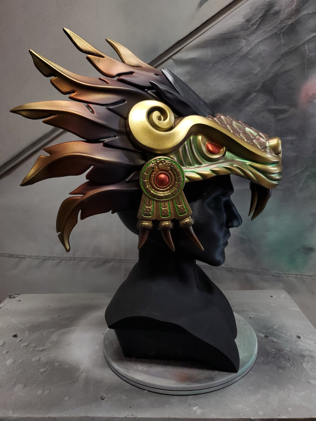 Hand‑sculpted Quetzalcóatl warrior mask with metallic gold, green, and violet detailing displayed on a black mannequin head