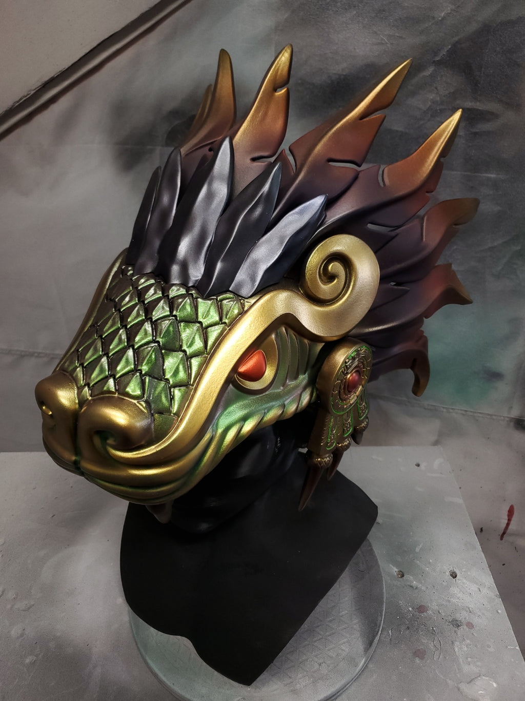 Hand‑sculpted Quetzalcóatl warrior mask with metallic gold, green, and violet detailing displayed on a black mannequin head