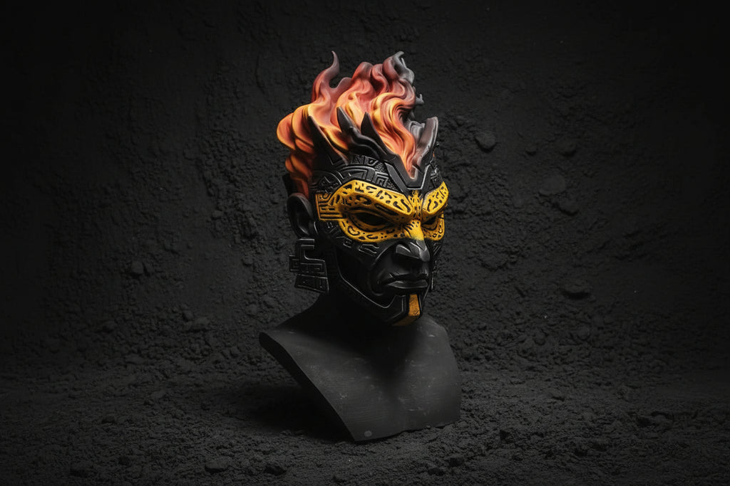 A black and gold fantasy mask with orange and yellow flames rising from the top, displayed on a black stand over a round base against a gray backdrop.