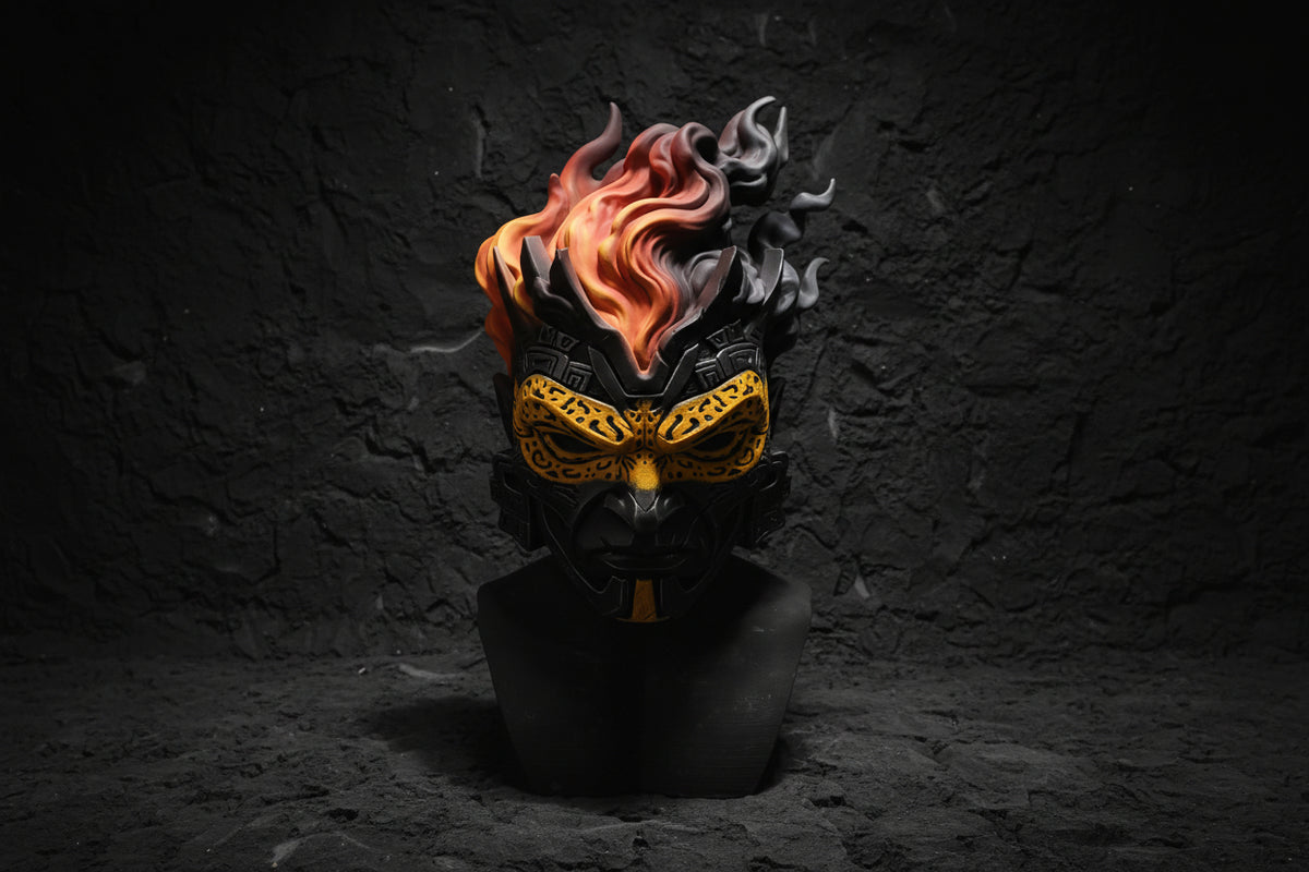A fantasy monster mask sculpture with black and gold facial details and vibrant orange and red flames rising from the top, displayed on a black stand against a gray backdrop.