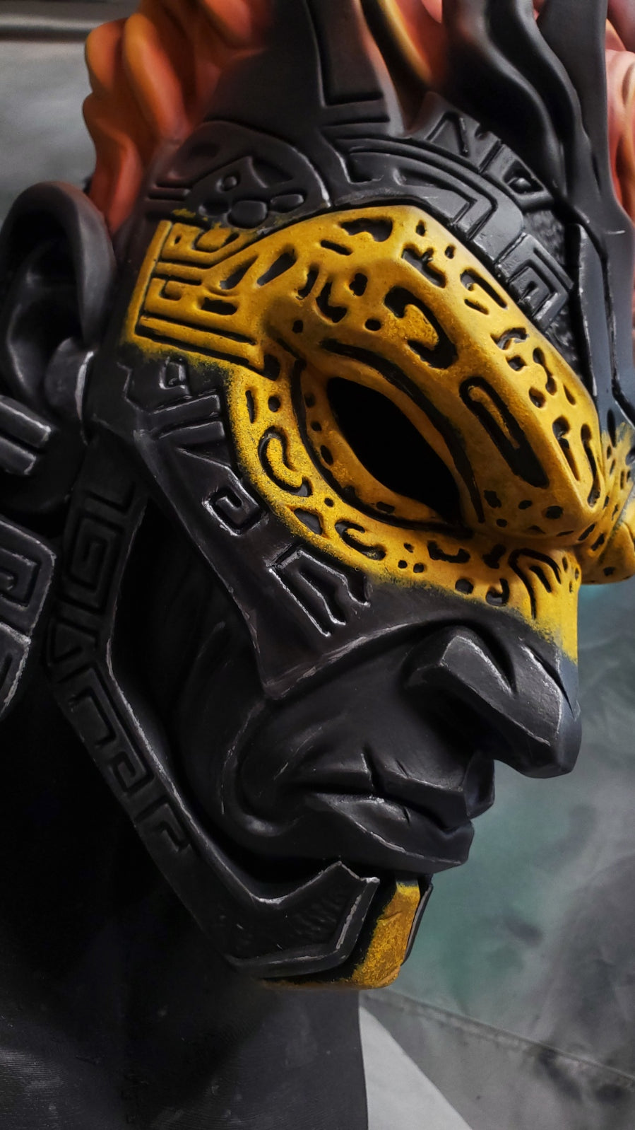 Close-up of a sculpted fantasy mask with a black base and a prominent yellow patterned eye section, shown against a dark blurred background.