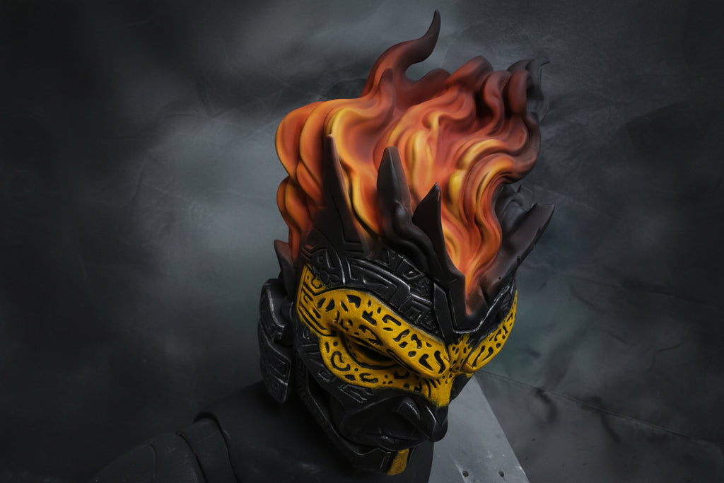 A close-up of a stylized fantasy helmet or mask with black angular armor, a bright yellow patterned lower face, and vivid orange-red flames rising from the top against a dark gray background.