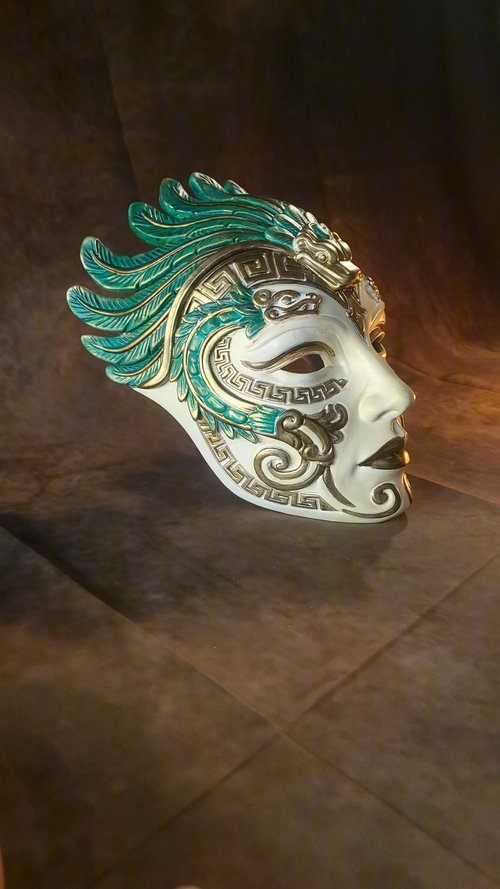 An ornate white masquerade mask with turquoise green feather-like detailing and gold accents, shown on a dark brown textured background.