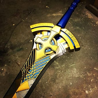 A decorative fantasy sword with a blue handle, gold and yellow guard, and black and blue patterned blade, photographed on a dark textured floor.