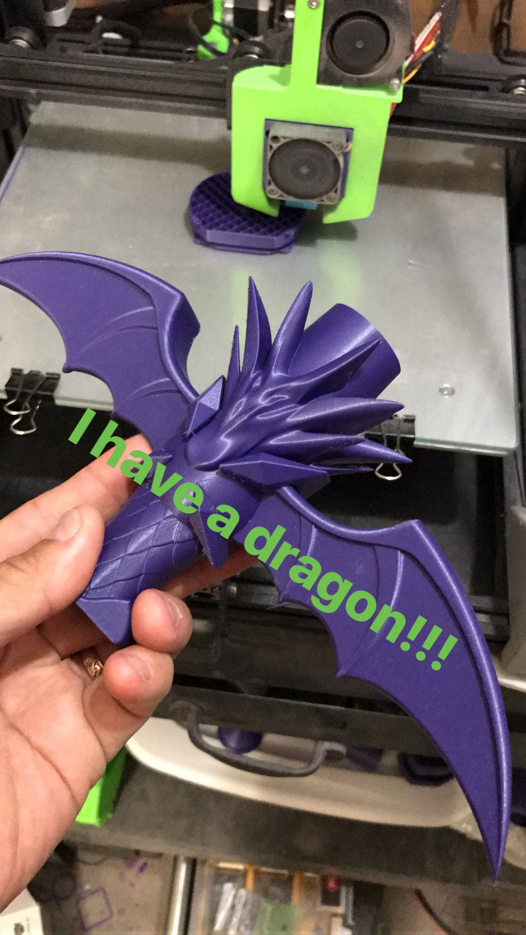 Purple dragon-shaped object with 'I have a dragon!!!' text held by a hand in front of a 3D printer.