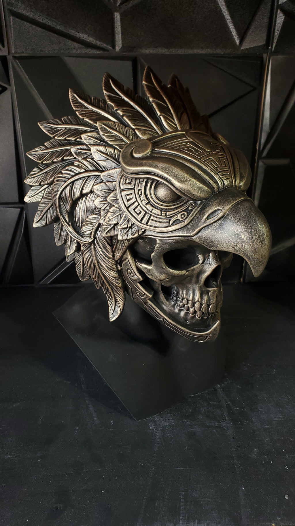 An ornate metallic bird skull mask with feather details and carved geometric patterns, displayed on a black stand against a dark textured backdrop.