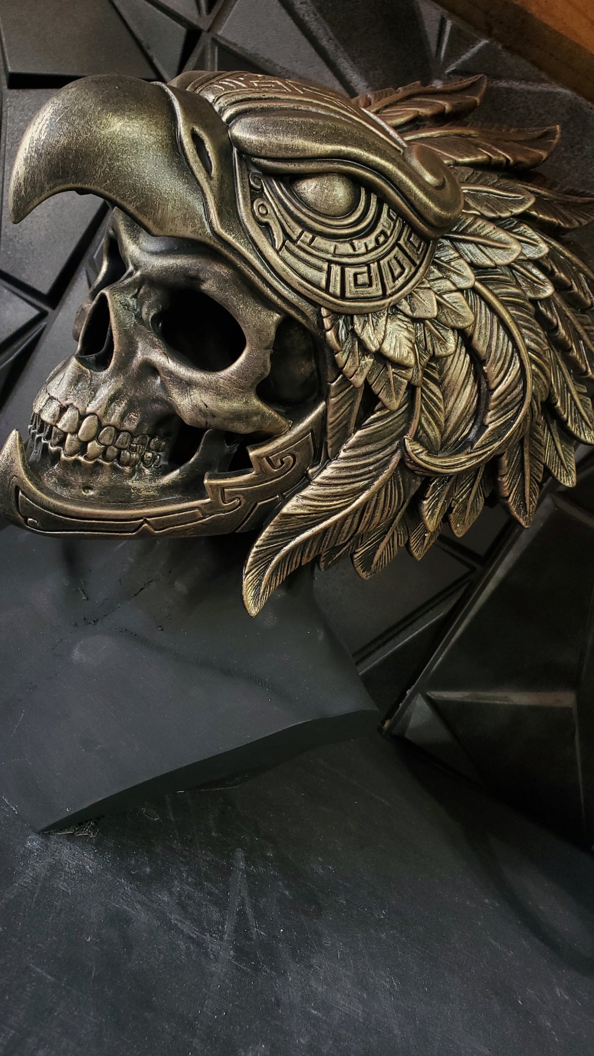 An ornate metallic eagle skull mask with feather and eye details, shown close up against a dark textured background.