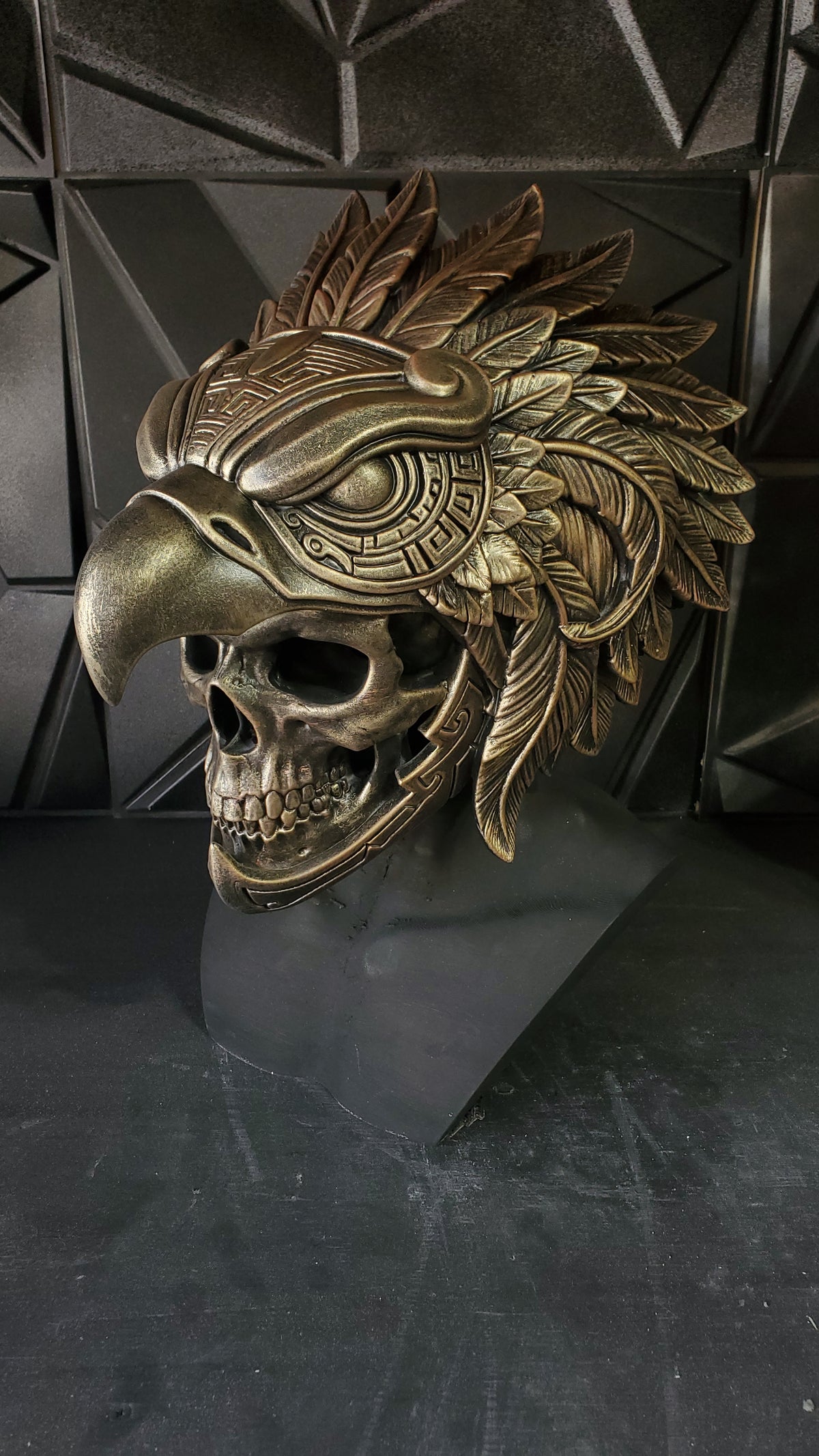 An ornate metallic eagle-and-skeleton mask displayed on a dark pedestal against a textured black geometric wall. The mask features a detailed beak, skull face, and layered feather motifs with engraved patterns.