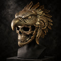 An ornate metallic skull mask with a large bird-like crest and feather details, displayed on a black stand against a dark geometric wall.
