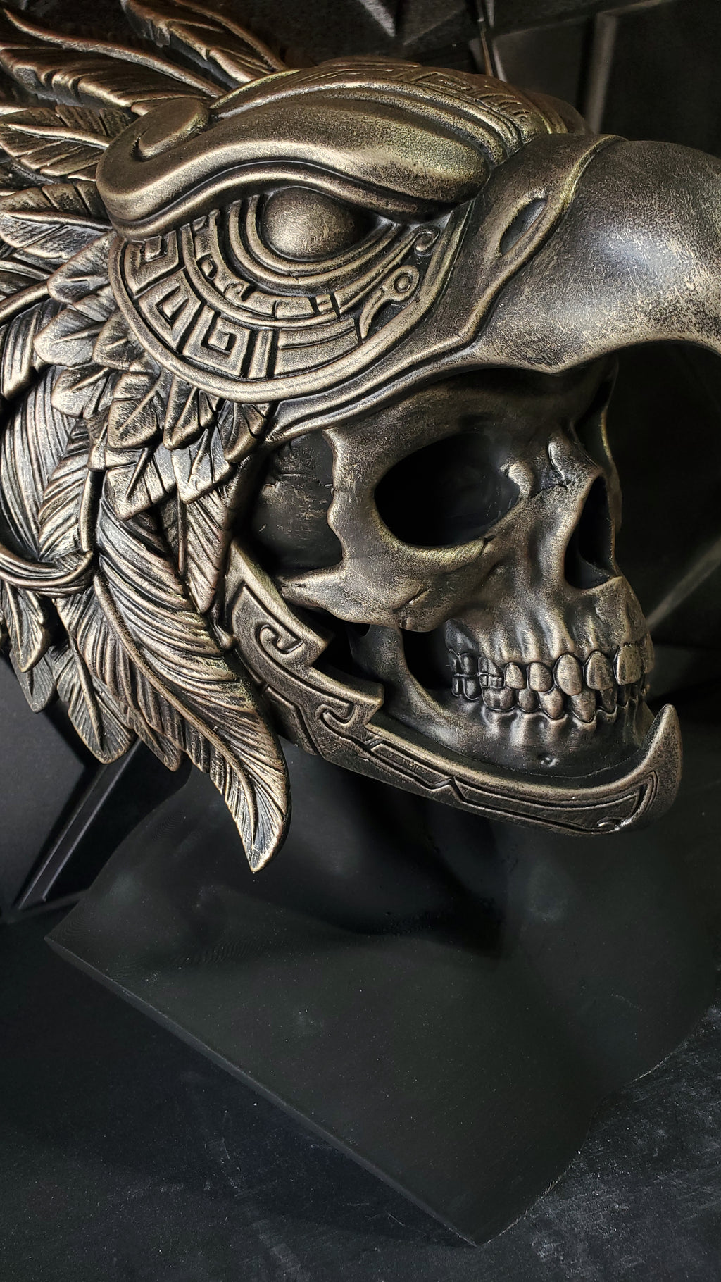 An ornate metallic sculpture combining a human skull with a bird of prey head and feather details, shown on a dark display base against a dark background.