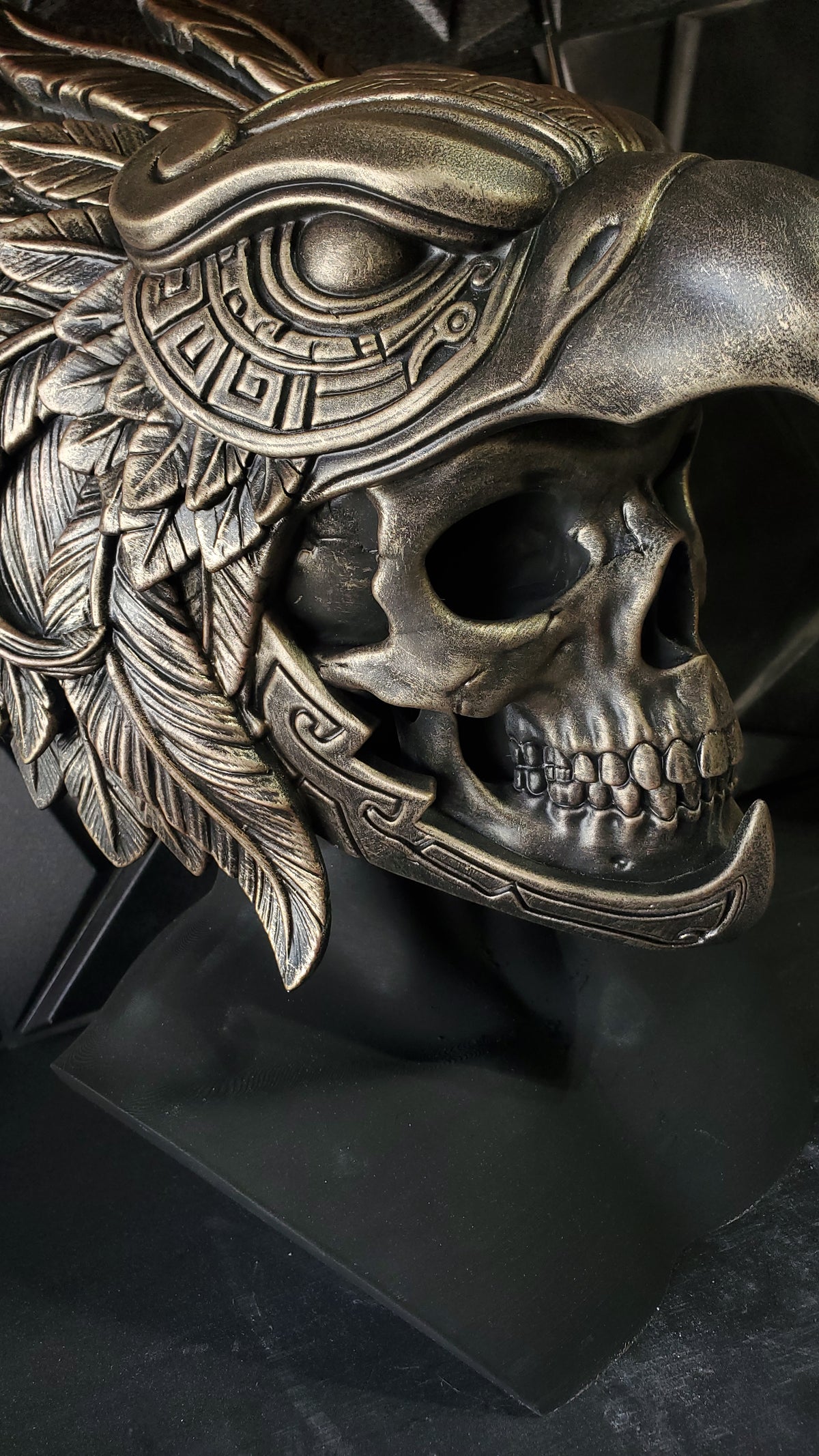An ornate metallic sculpture combining a human skull with a bird of prey head and feather details, shown on a dark display base against a dark background.